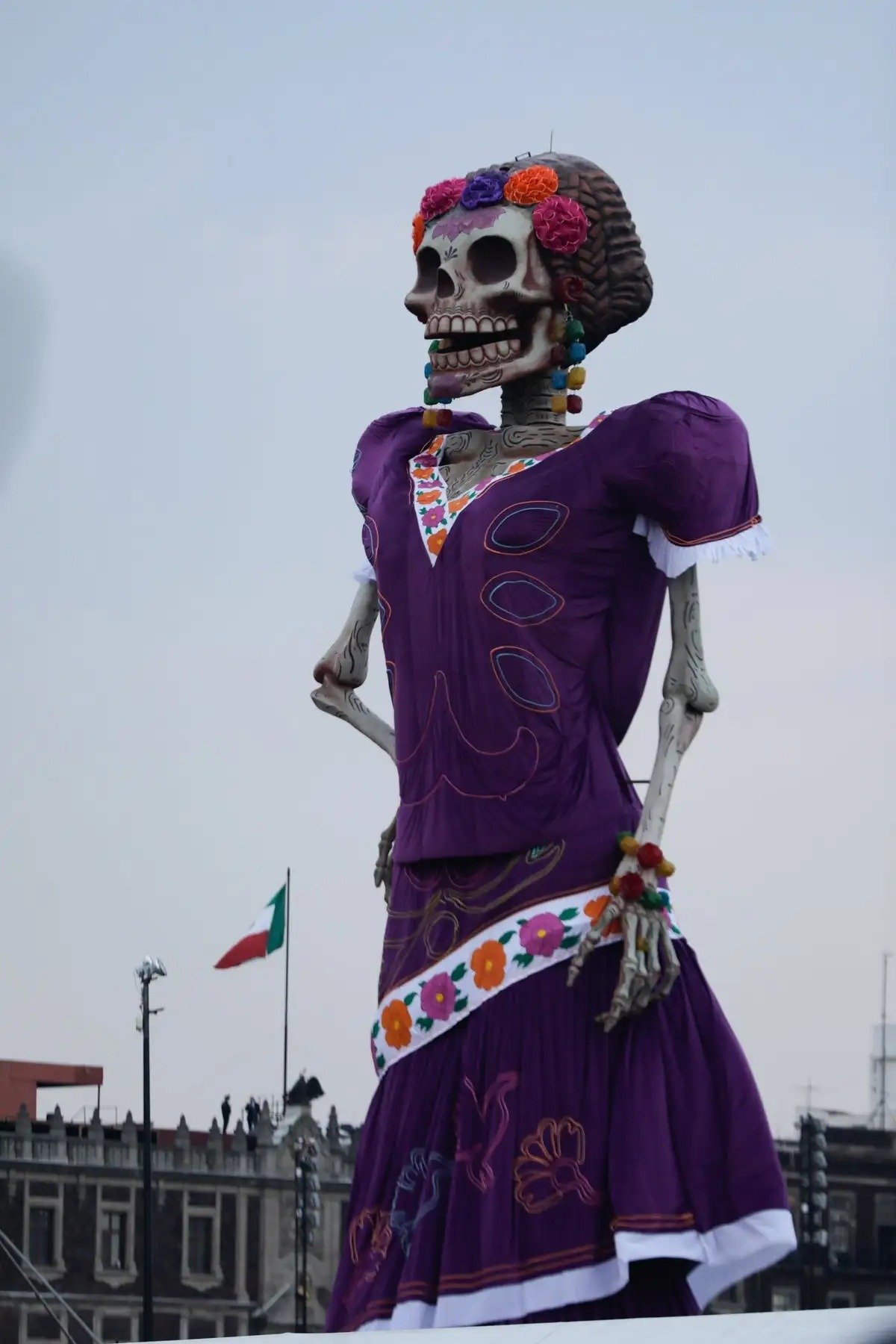 Close-up of the giant catrina in a purple dress and flower crown, with the Mexican flag in the background