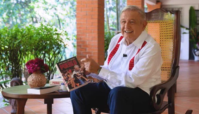 López Obrador, seated on a porch, holds up a copy of his book Pueblo.