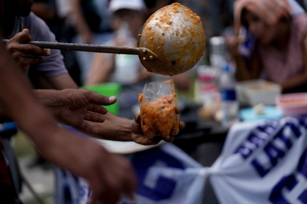 A hand ladles food into a plastic bag held out by another person at a community food distribution.
