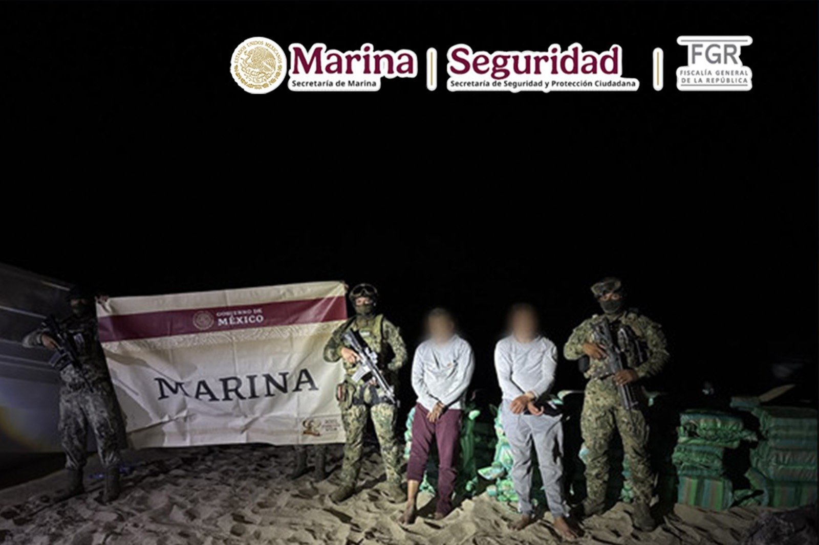 Navy personnel stand on a beach at night with two detainees and seized packages