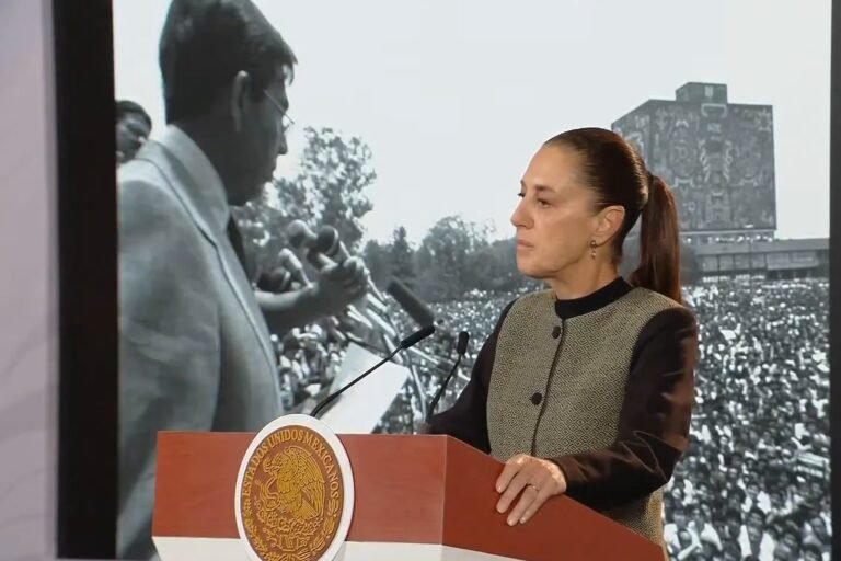 President Claudia Sheinbaum, in profile at the podium, in front of a black-and-white photo of Cuauhtémoc Cárdenas addressing a huge crowd at UNAM